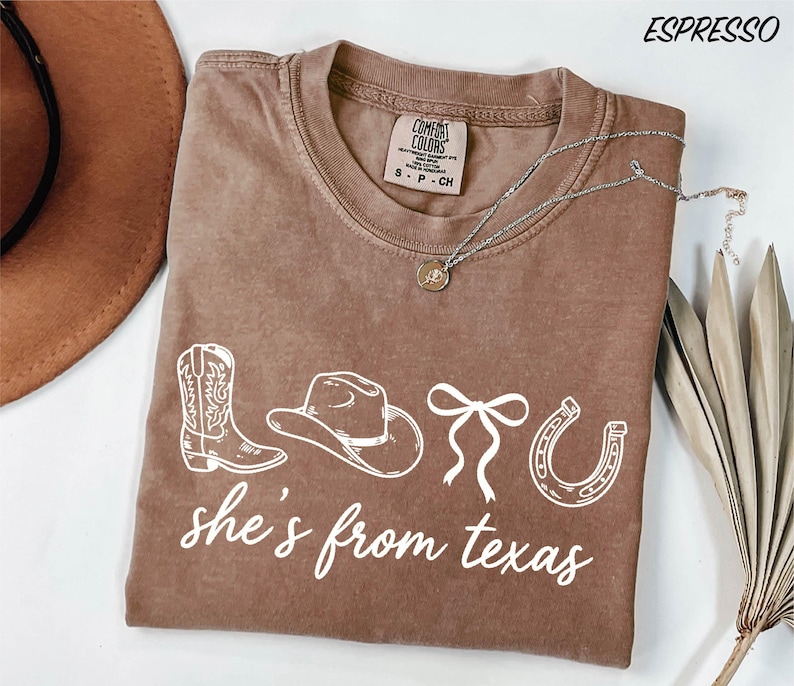 a brown t-shirt with the text "she's from texas" printed on it, along with various western-themed icons such as a cowboy hat, a boot, and a horseshoe. The t-shirt is laid out on a white surface, and there is a brown hat and some dried leaves nearby.