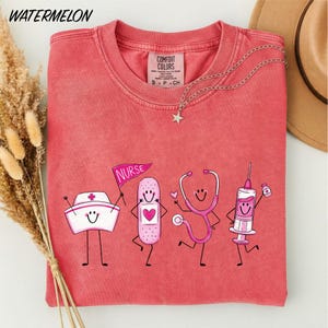 A pink t-shirt with a design of four cartoon nurse characters, including a nurse holding a stethoscope, a nurse holding a syringe, and a nurse holding a bandage, along with a star-shaped charm.