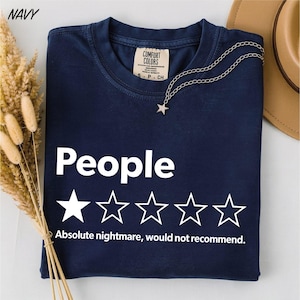 May include: A navy blue t-shirt featuring the white text "People" above a five-star rating graphic where only the first star is filled in. Below the stars, the white text reads "Absolute nightmare, would not recommend." A silver star pendant necklace rests on the collar.