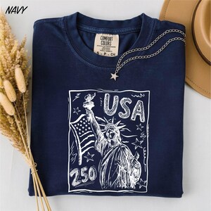 A navy blue t-shirt with a graphic design featuring the Statue of Liberty and the text "USA 250" printed on it, along with a necklace and some dried flowers.