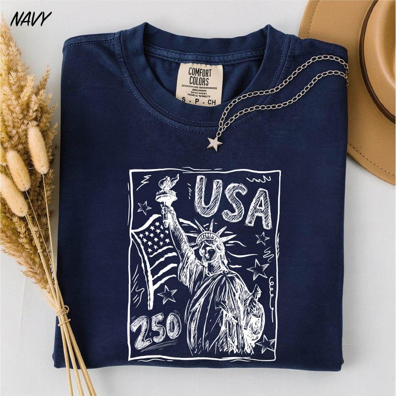 A navy blue t-shirt with a graphic design featuring the Statue of Liberty and the text "USA 250" printed on it, along with a necklace and some dried flowers.