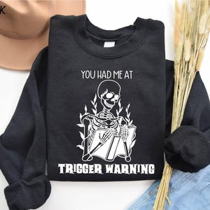 May include: Black sweatshirt with a white skeleton reading a book and the text "You had me at trigger warning".