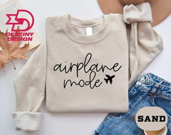 Puede incluir: Una sudadera de color arena con la frase "airplane mode" en fuente negra, acompañada de un pequeño gráfico de avión. La sudadera está doblada, con unos vaqueros azules y un sombrero en el fondo.