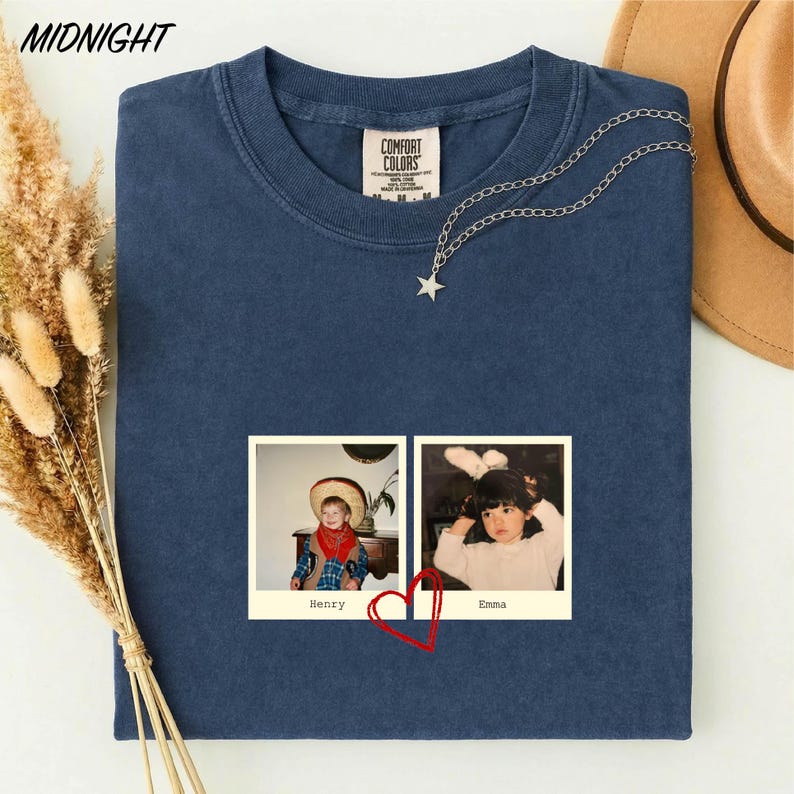 a blue t-shirt with a photo of two children, one wearing a cowboy hat and the other wearing a white shirt, along with a heart symbol. The t-shirt is placed on a white surface, and there is a brown hat and some dried flowers in the background.