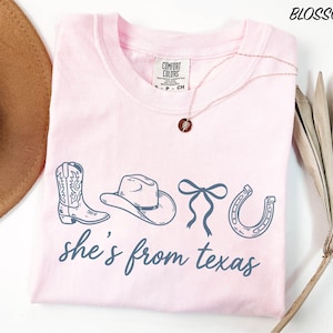 A pink t-shirt with the text "she's from texas" printed on it, along with illustrations of cowboy boots, a hat, and a horseshoe. The t-shirt is laid out on a white surface, and there is a brown hat and a plant in the background.