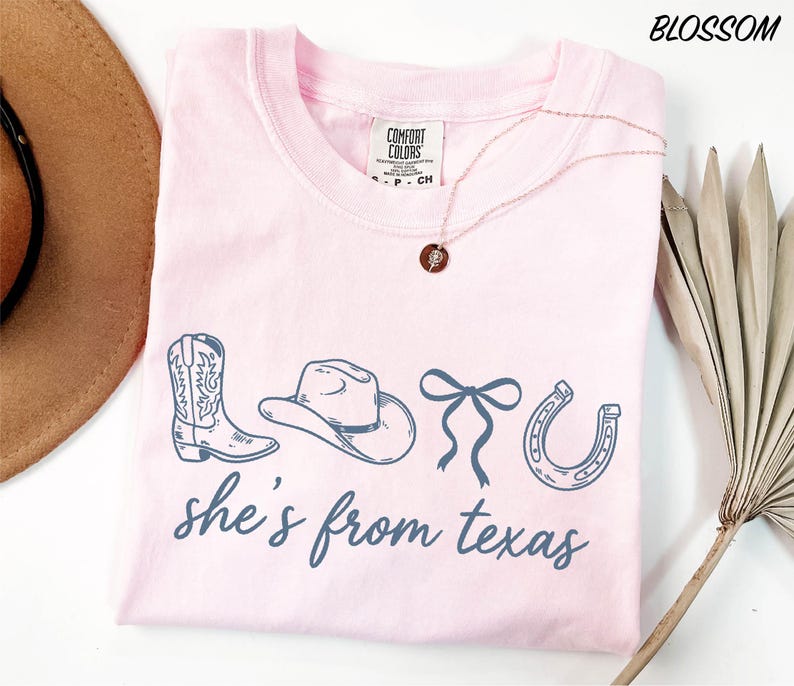 A pink t-shirt with the text "she's from texas" printed on it, along with illustrations of cowboy boots, a hat, and a horseshoe. The t-shirt is laid out on a white surface, and there is a brown hat and a plant in the background.