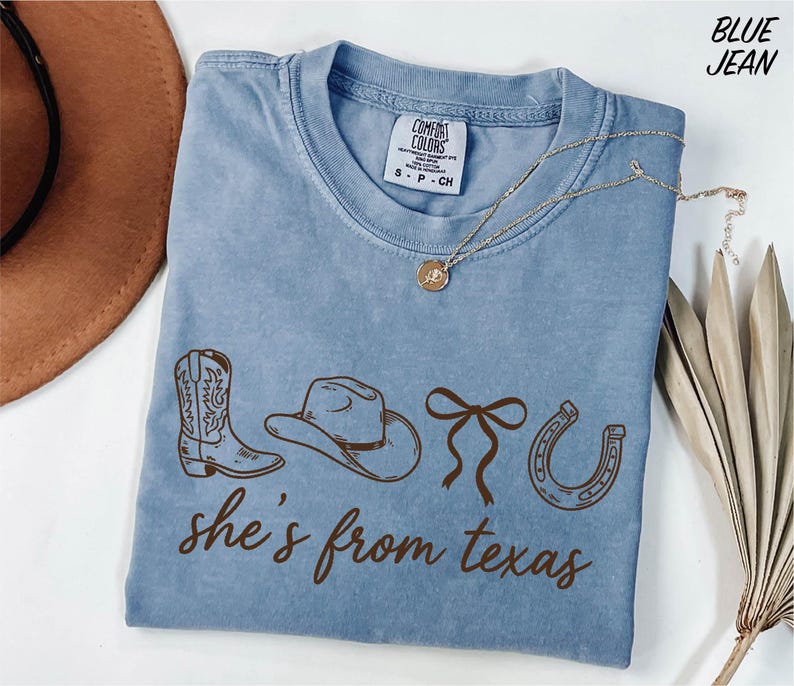 A blue t-shirt with the text "she's from texas" printed on it, along with illustrations of cowboy boots, a hat, and a horseshoe. The t-shirt is laid out on a white surface, and there is a brown hat and some dried leaves nearby.