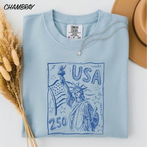 A light blue t-shirt with a graphic design featuring the Statue of Liberty and the text "USA 250" printed on it, along with a star pendant. The t-shirt is placed on a white surface, and there are some dried flowers and a brown hat nearby.