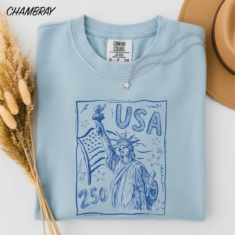 A light blue t-shirt with a graphic design featuring the Statue of Liberty and the text "USA 250" printed on it, along with a star pendant. The t-shirt is placed on a white surface, and there are some dried flowers and a brown hat nearby.