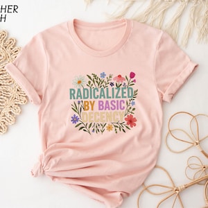 May include: Heather peach-colored t-shirt with the text "RADICALIZED BY BASIC DECENCY" in a floral design. The shirt has short sleeves and a round neckline. The text is in various colors, including green, purple, and yellow.