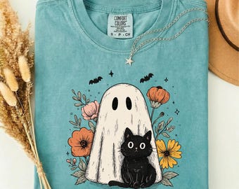 Puede incluir: Una camiseta verde espuma de mar con un gráfico de un fantasma blanco y un gato negro sentados juntos, rodeados de flores naranjas y amarillas y pequeños murciélagos negros. La camiseta tiene cuello redondo y una etiqueta de Comfort Colors.