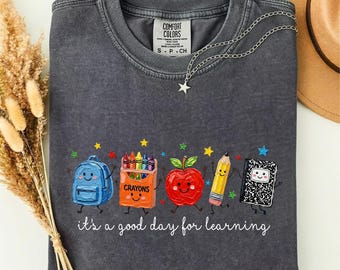 Puede incluir: Una camiseta de cuello redondo gris carbón con un gráfico de una mochila azul, una caja de ceras naranja, una manzana roja, un lápiz amarillo y un cuaderno negro con caras. El texto dice, it's a good day for learning.