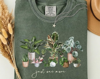 May include: A sage green crewneck t-shirt featuring a graphic print of various potted houseplants and the text "just one more" in white script. The shirt has a ribbed collar and a tag that reads Comfort Colors.