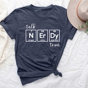 May include: Dark gray t-shirt with the phrase "talk NERDY to me" in white. The word "NERDY" is spelled out using the periodic table symbols for nitrogen, erbium, and dysprosium. The shirt is knotted at the bottom.