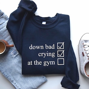 May include: Navy blue sweatshirt with white text that reads "down bad crying at the gym" with checkboxes next to each item. The checkboxes next to "down bad" and "crying" are checked.