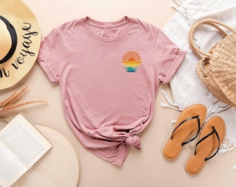 May include: A dusty rose t-shirt with a small, colorful sunset graphic. The shirt is styled with a straw hat, sandals, a woven bag, and an open book. The hat has the words "en voyage" printed on it.