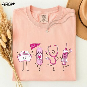 A pink t-shirt with a graphic design of four cartoon nurse characters, including a nurse, stethoscope, syringe, and bandage, along with the text "NURSE" and "CONFIDENCE COLOR".