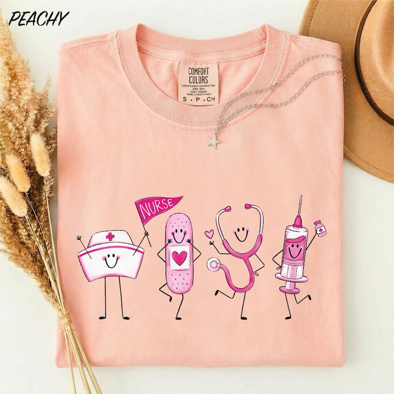 A pink t-shirt with a graphic design of four cartoon nurse characters, including a nurse, stethoscope, syringe, and bandage, along with the text "NURSE" and "CONFIDENCE COLOR".