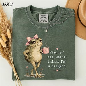 May include: A sage green t-shirt featuring a graphic of a toad wearing a pink bow, holding a pink mug, and the text: first of all, Jesus thinks i'm a delight. The shirt has a crew neckline and a Comfort Colors brand label on the inner collar.