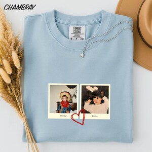 A light blue t-shirt with a photo of two children, one wearing a cowboy hat and the other a white dress, printed on it. The photo is accompanied by a heart symbol.