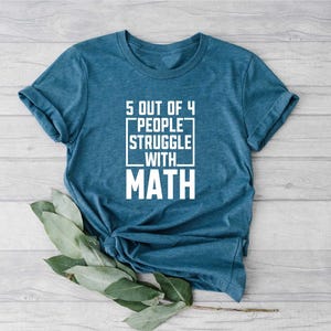 May include: A teal-colored t-shirt with the text "5 OUT OF 4 PEOPLE STRUGGLE WITH MATH" in white block letters. The text is centered and enclosed in a white rectangular border. The shirt is folded and lies on a light wood surface.