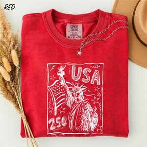A red t-shirt with a graphic design of the Statue of Liberty and the text "USA 250" printed on it, along with a star charm.