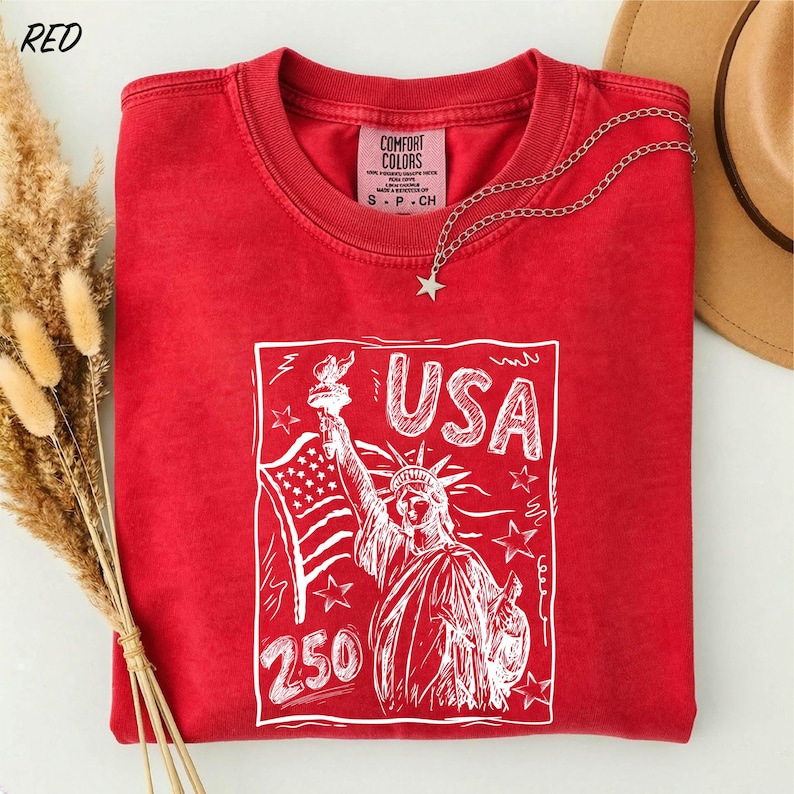 A red t-shirt with a graphic design of the Statue of Liberty and the text "USA 250" printed on it, along with a star charm.