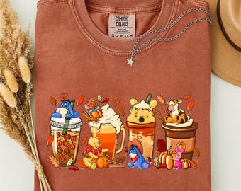 Może przedstawiać: T-shirt z okrągłym dekoltem w kolorze rdzawym z nadrukiem postaci z Kubusia Puchatka, jesiennymi napojami, dyniami i liśćmi. Koszulka posiada metkę Comfort Colors przy wewnętrznym dekolcie oraz srebrny naszyjnik z zawieszką w kształcie gwiazdy.