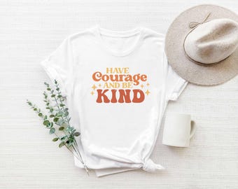 May include: White t-shirt with the text "HAVE Courage AND BE KIND" in orange and rust-colored retro font. The shirt is styled with a beige hat, a white mug, and sprigs of greenery on a white surface.