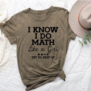 May include: Olive green t-shirt with the text "I KNOW I DO MATH Like a Girl TRY TO KEEP UP" in black. The shirt is knotted at the bottom and is next to a beige hat with a brown band and some greenery.