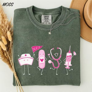 a green t-shirt with a graphic design of four cartoon-style medical professionals, including a nurse, a stethoscope, a syringe, and a pill bottle, all depicted in a playful and colorful manner.