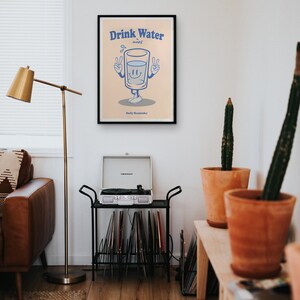 K&ouml;nnte beinhalten: Ein schwarzer gerahmter Poster mit einer Cartoon-Illustration eines Glases Wasser mit Armen und Beinen. Das Glas l&auml;chelt und zeigt mit beiden H&auml;nden das Peace-Zeichen. Der Text auf dem Poster lautet "Drink Water A Daily Reminder".