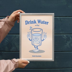 K&ouml;nnte beinhalten: Ein gerahmtes Poster mit einem hellrosa Hintergrund und einer Cartoon-Illustration eines Glases Wasser mit einem l&auml;chelnden Gesicht. Der Text auf dem Poster lautet "Drink Water More Daily Reminder".