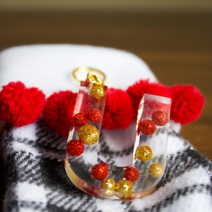 May include: A clear resin letter U ornament with red and gold glitter balls inside. The ornament is hanging from a gold chain and is resting on a white and black plaid fabric.