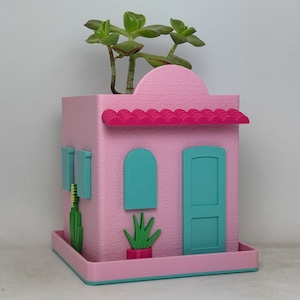 May include: A pink and turquoise 3D printed planter shaped like a house with a green succulent plant inside. The planter has a red roof, two windows, and a door.
