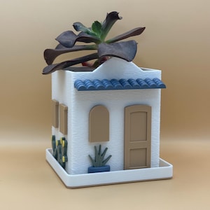 May include: White ceramic planter shaped like a small house with a blue tiled roof. The planter has a brown door and windows, and a small green plant is growing out of the top.