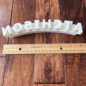 May include: White plastic letters spelling out the word "ATCHISON" in a curved shape. The letters are on a wooden surface.
