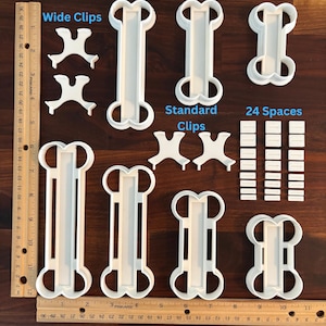 May include: A set of white plastic cookie cutters in the shape of bones. The cutters are in four sizes: 15 cm, 12.5 cm, 10 cm, and 7.5 cm. The set also includes two pairs of white plastic clips and 24 white plastic spacers.