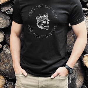 Peut inclure: Un t-shirt noir avec un graphique blanc d'un cr&acirc;ne portant une couronne. Le texte sur le t-shirt est "I only like disc golf and about 3 people."