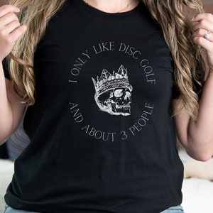 Peut inclure: T-shirt noir avec un graphique blanc d'un cr&acirc;ne portant une couronne. Le graphique dit "I only like disc golf and about 3 people".
