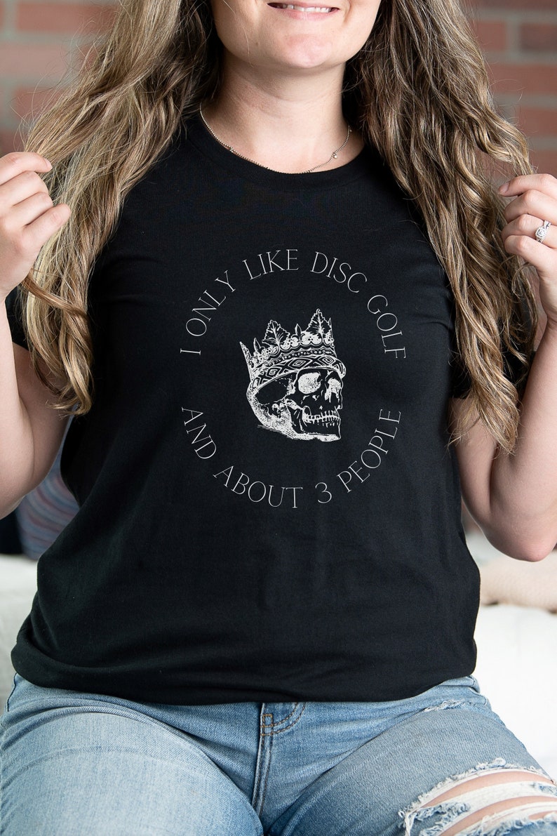 Peut inclure: T-shirt noir avec un graphique blanc d'un cr&acirc;ne portant une couronne. Le graphique dit "I only like disc golf and about 3 people".