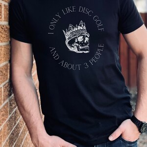 Peut inclure: Un t-shirt noir avec un motif blanc d'un cr&acirc;ne portant une couronne. Le texte sur le t-shirt est "I only like disc golf and about 3 people."