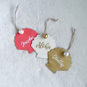 May include: Three wooden seashell-shaped place cards with white pearl accents. The place cards are painted in pink, white, and gold. The names "Jourdan", "Aloha", and "Shelly" are written in white on the place cards.