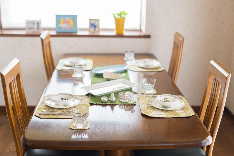Puede incluir: Un juego de comedor de madera con cuatro sillas, cubiertos y manteles individuales verdes. La mesa est&aacute; puesta con platos blancos, cuencos, vasos y cubiertos. Tambi&eacute;n hay dos peque&ntilde;os saleros y pimenteros de plata en la mesa.