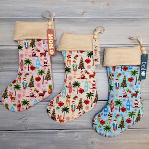 May include: Three Christmas stockings with a Hawaiian theme. The stockings are decorated with palm trees, surfboards, and Santa Claus wearing a lei. The stockings are labeled "Mom", "Leilani", and "Dad".