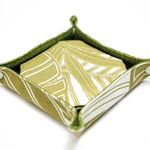 May include: A small, square-shaped, green fabric tray with a white and green leaf pattern. The tray is folded up and has a white background.