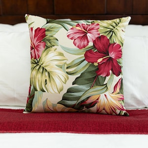 May include: Square decorative pillow with a tropical floral pattern. The pillow features large red hibiscus flowers, green leaves, and yellow accents on a cream background. The pillow is placed on a red bed runner.