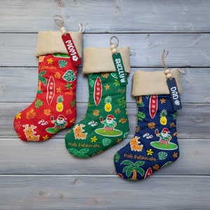 Puede incluir: Tres calcetines de Navidad con temática hawaiana. Los calcetines son rojo, verde y azul con un diseño de Papá Noel surfeando en una tabla de surf. Cada calcetín tiene una etiqueta de madera personalizada con los nombres "Mamá", "Shelly" y "Papá".