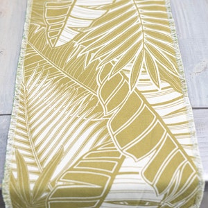 May include: A green and white table runner with a tropical leaf pattern. The runner has a frayed edge.