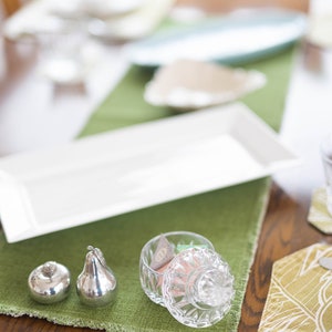 Puede incluir: Un plato rectangular blanco, dos saleros y pimenteros de plata en forma de pera y un frasco de vidrio con tapa lleno de caramelos de colores sobre un mantel verde.